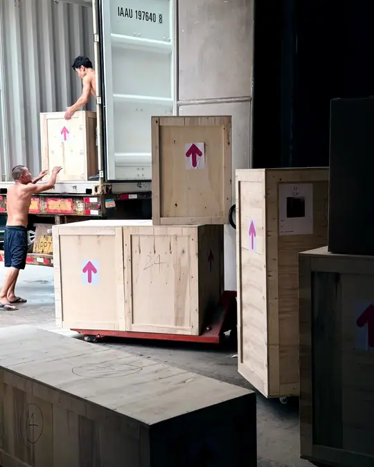 Two warehouse workers guiding a numbered wooden shipping crate into a container, with more crated furniture staged on pallets alongside