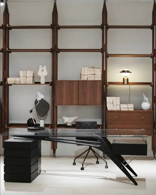 A home office styled with a black cantilever desk, an office chair and a wall of walnut and glass shelving holding books and ceramics