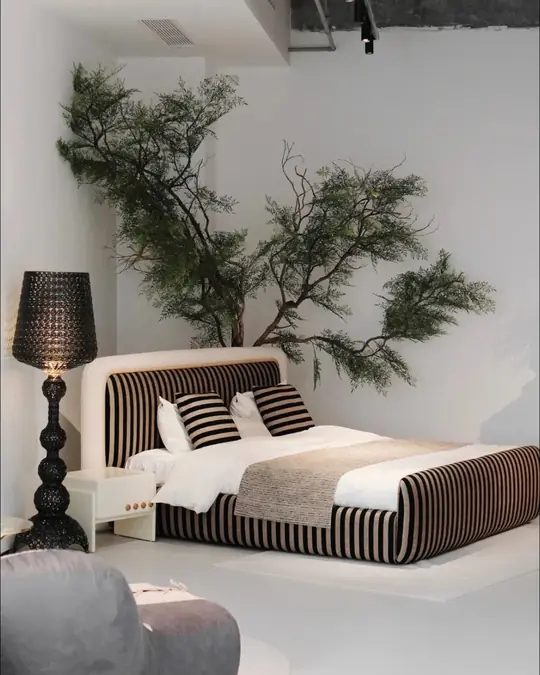 A black and white striped fabric daybed with a grey boucle ottoman and a turned black floor lamp beside an indoor tree