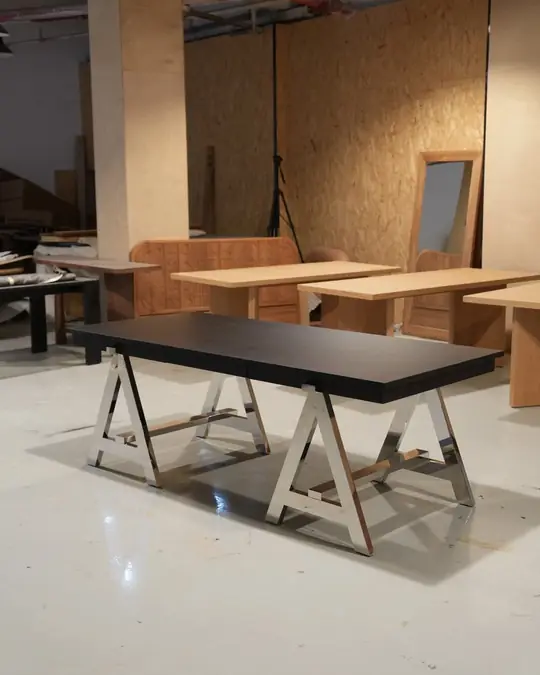 A dining table with a black top on pale timber trestle legs, shown in a showroom alongside other tables