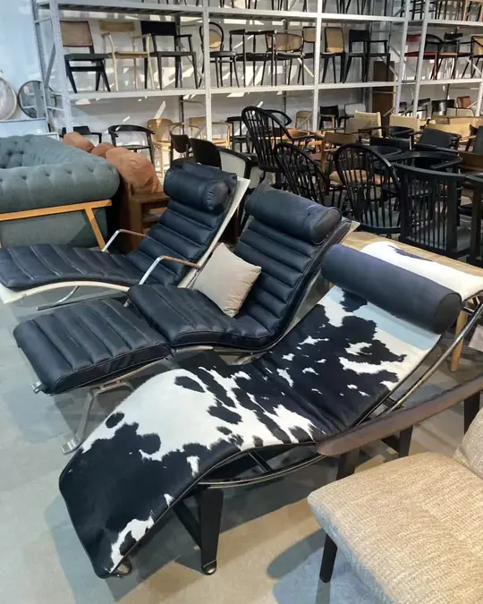 A black leather reclining lounge chair with a cowhide-patterned throw, beside a grey button-tufted sofa, in a showroom of stacked chairs