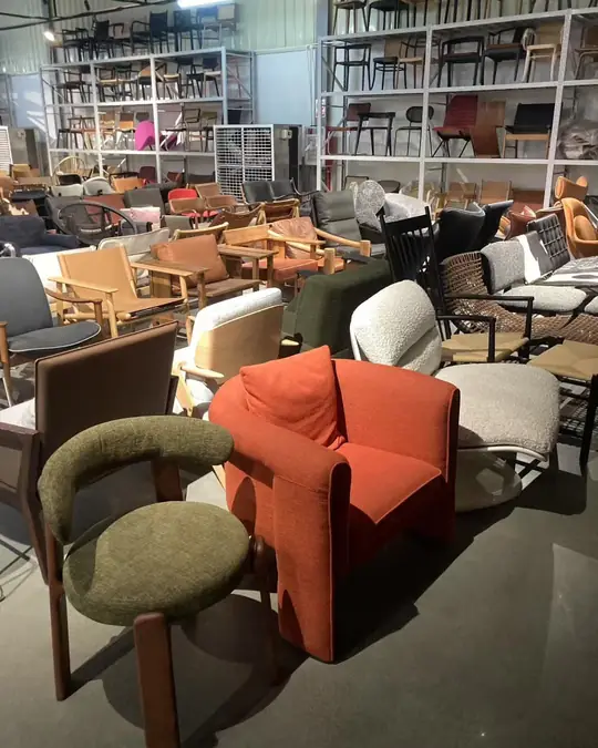 A rust-orange boucle armchair and an olive green wool chair in the foreground of a showroom lined with racking of dining chairs