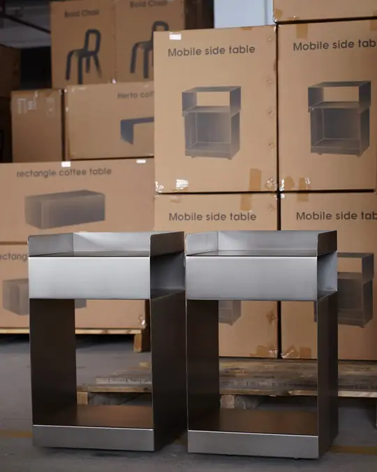 Two grey metal side tables standing in front of stacked cartons printed with their product photograph