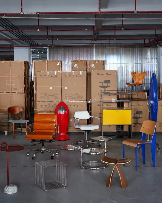 A warehouse corner of stacked shipping cartons with orange, yellow and red designer chairs set out in front of them